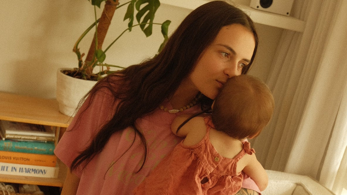 Woman holding a child in a living room with a plant and books in the background